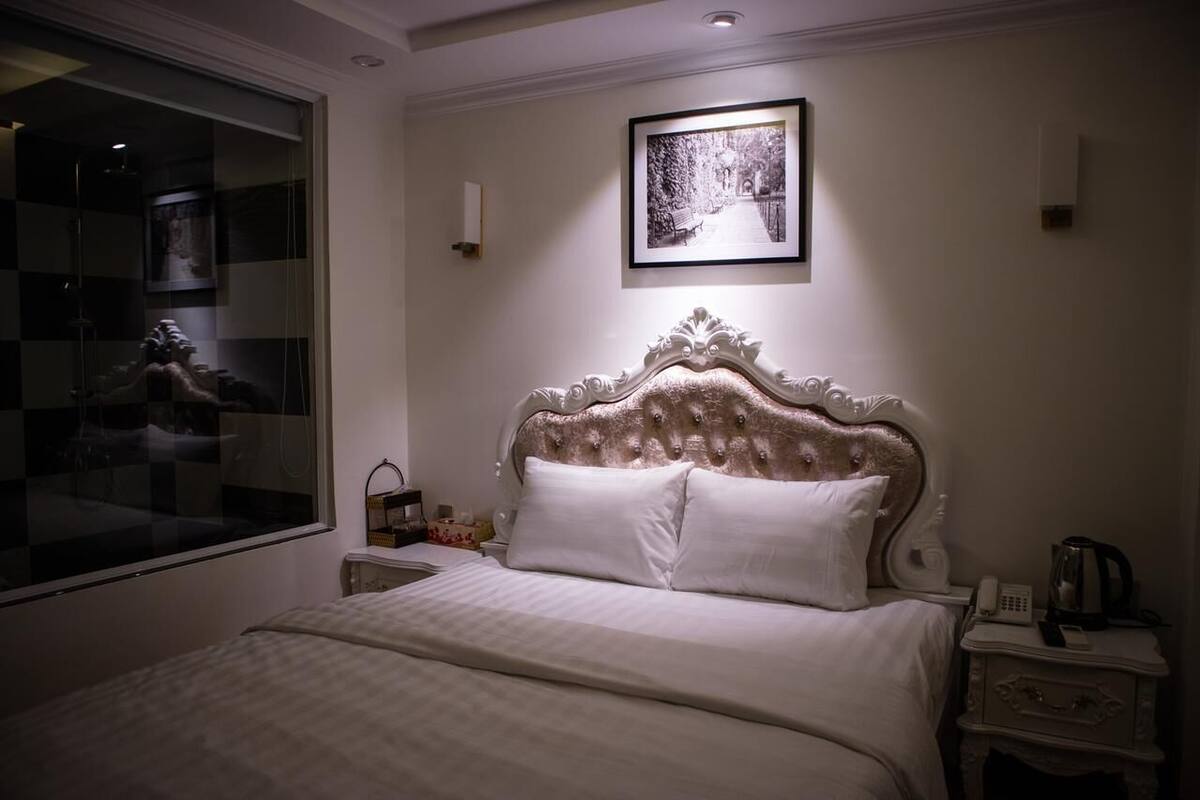 Une chambre à coucher avec un grand lit, une table de chevet avec une lampe, un tableau encadré au mur et une fenêtre donnant sur un immeuble.
