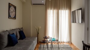 A modern living room with a sofa, a small table with decorative items, a vase with flowers, and a large window with curtains. - Acropolis Grand Apartment (Athina)