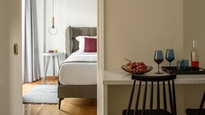 A hotel room with a bed, bedside table, and a small dining area with a bowl of fruit and wine glasses. - Acropolis Grand Apartment (Athina)