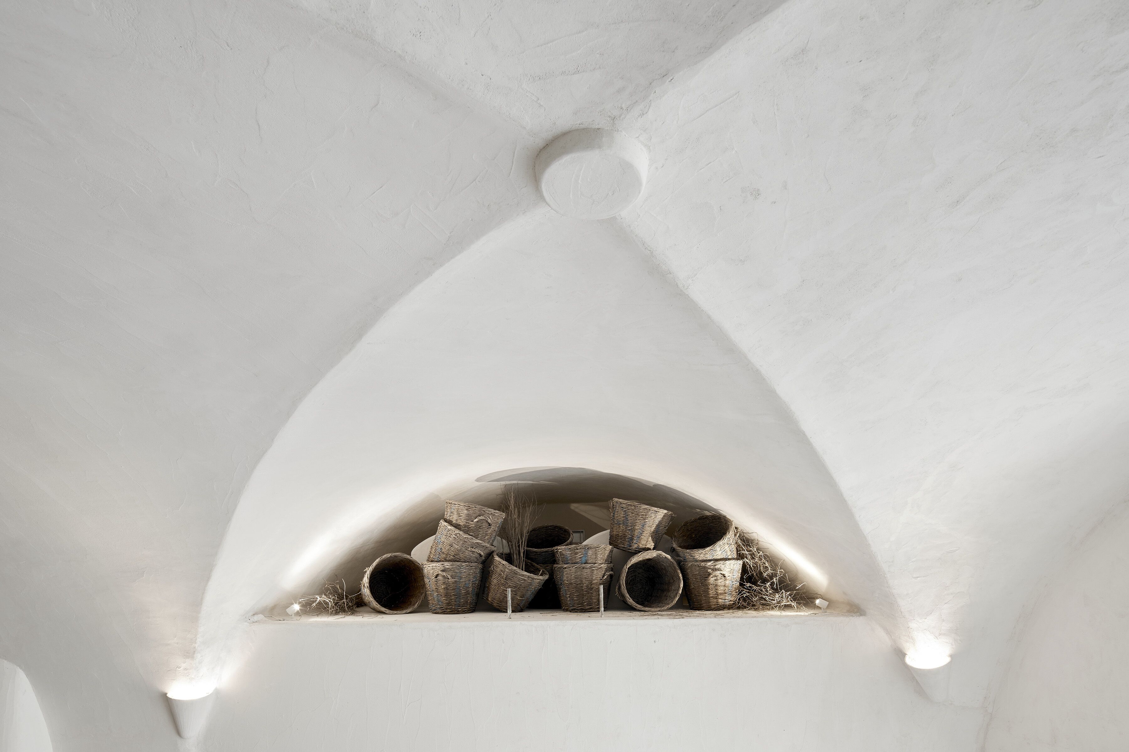 A white arched alcove with a small window, containing several wicker baskets and a straw broom.