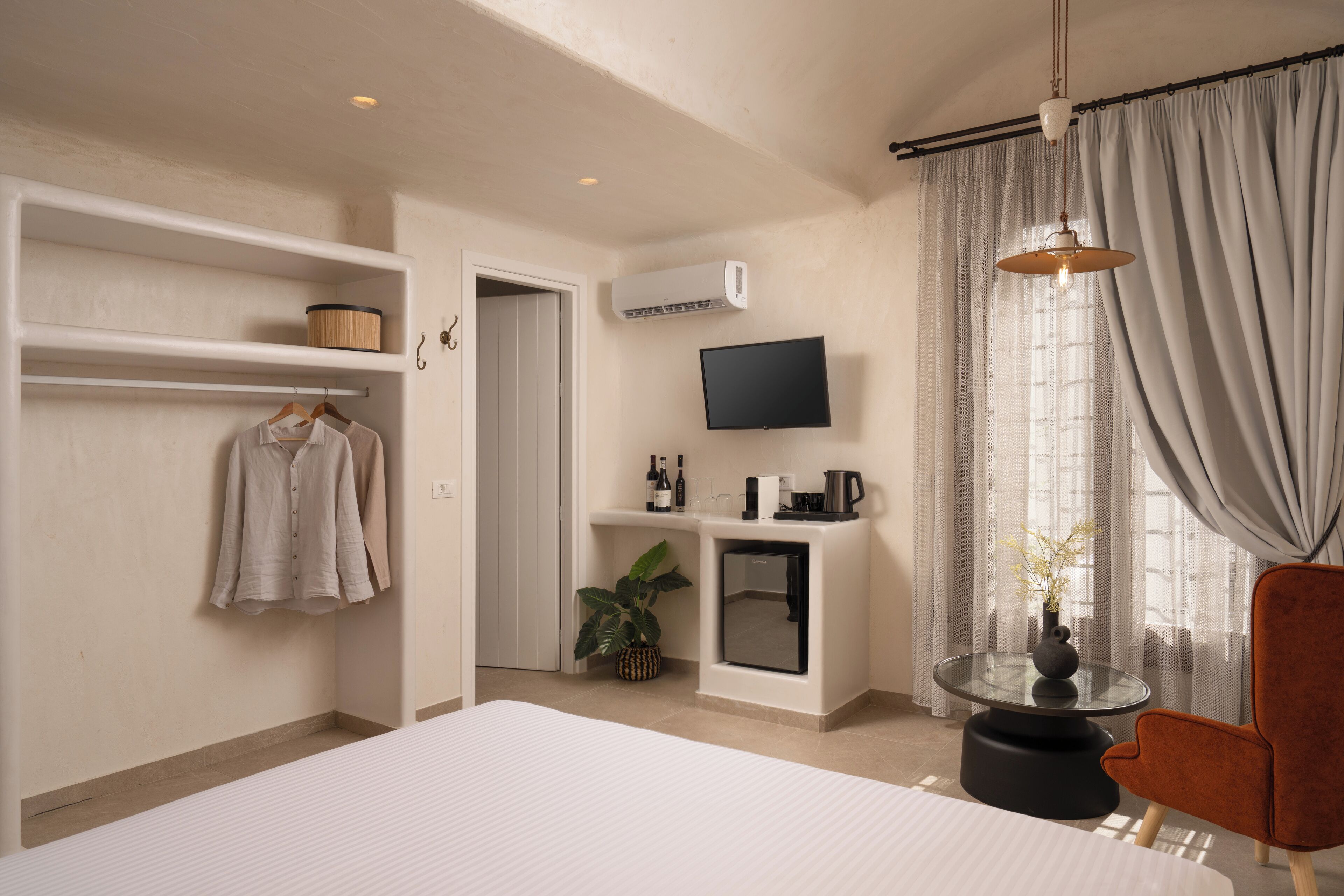 Habitación de hotel con cama, zona de estar con una silla, televisor montado en la pared, una mesita con un florero y una planta.