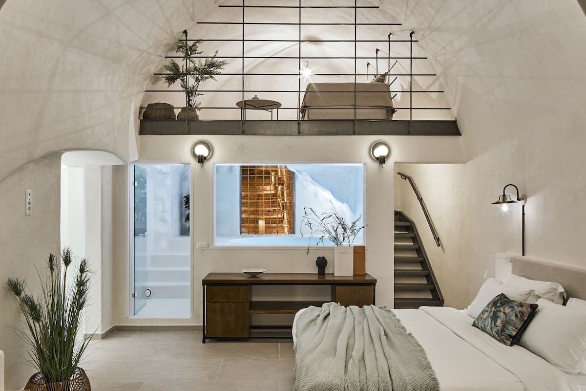 Een ruime loft slaapkamer met een hoge plafond, een groot bed, een bureau en een trap naar een hoger niveau.