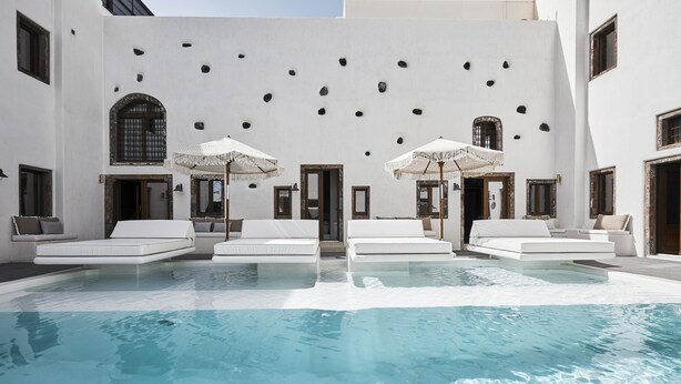 Zona junto a la piscina con sillas reclinables blancas y sombrillas, una piscina cristalina y un edificio con fachada blanca y ventanas de arco.