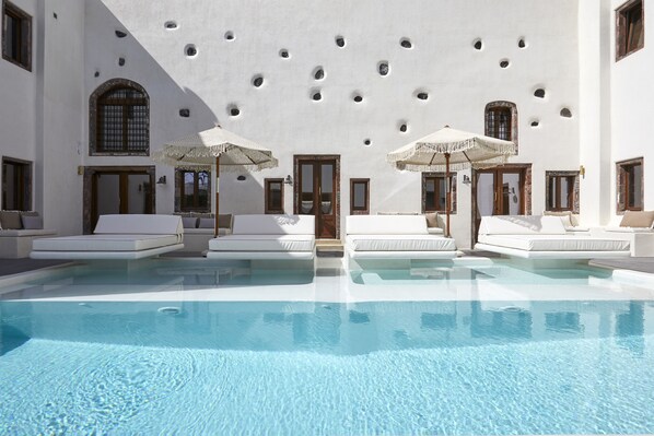 Un área de piscina con sillones blancos y sombrillas, una piscina cristalina y un edificio con ventanas arqueadas y techo decorado.
