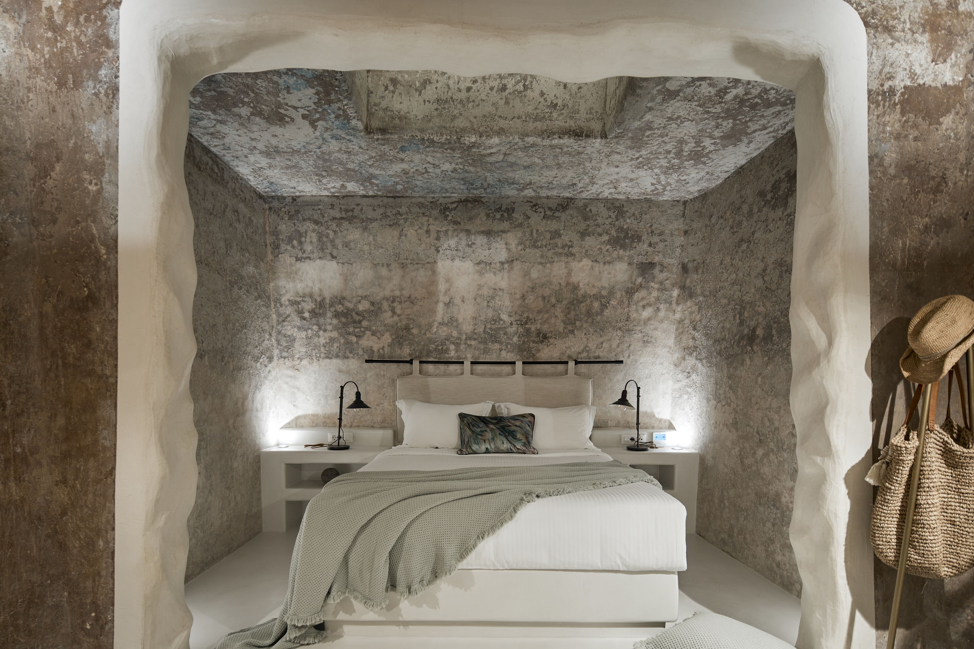 Un dormitorio con pared de piedra, una cama con cabecera blanca, dos mesitas de noche con lámparas y un bolso tejido colgado en la pared.