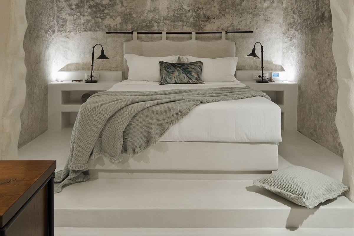 Een slaapkamer met een rustieke stenen muur, een bed met wit beddengoed, twee bedlampjes en een klein bureau met lamp.