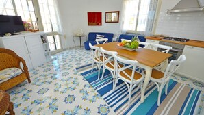 Un coin repas avec une table en bois et des chaises blanches, une cuisine équipée d’une cuisinière et un sol à motifs.