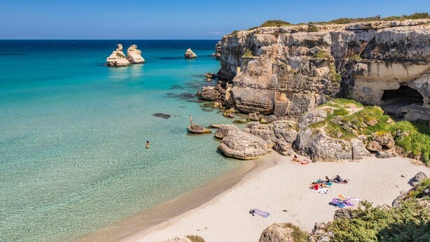 Une plage à l’eau turquoise et limpide, bordée de falaises rocheuses et d’une étendue de sable où les gens se détendent.