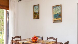A dining room with a wooden table set for four, two paintings on the wall, and a window with curtains.