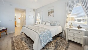 A bedroom with a bed, a nightstand, a chair, a window with curtains, a ceiling fan, and a door leading to another room.