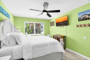 A bedroom with a bed, a ceiling fan, a window with blinds, a television, and a wall-mounted coat rack.