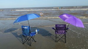 Deux chaises de plage, chacune munie d’un parasol, l’une bleue et l’autre violette, sur une plage de sable, avec l’océan en arrière-plan.