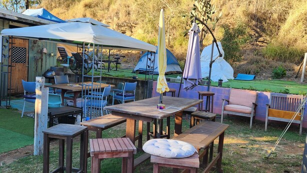 An outdoor seating area with wooden tables and chairs, a covered patio, and a tented accommodation in the background. - Meyenburg Unit 5, 2 Double Bed's 4 Pax (Mount Morgan)
