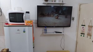 Ein gemütlicher Wohnbereich mit einem weißen Kühlschrank, einer Mikrowelle, einem an der Wand befestigten Fernseher, einem kleinen gelben Hocker und einem Esstisch mit Kaffeemaschine.
