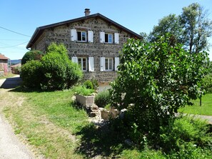 A stone house with white shutters, surrounded by lush greenery and a gravel path.