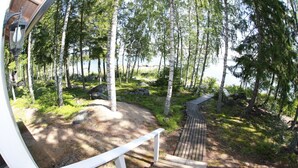 A wooden deck with a white railing, surrounded by a forested area with birch trees and a view of a lake. - Seaside Villa with jacuzzi and own beach (Maksamaa (Vöyri))