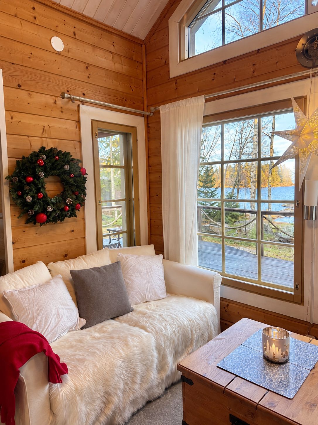 A cozy cabin interior with a wooden interior, a white sofa, a table with a candle, and a view of trees and water outside.