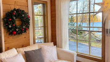 A cozy cabin interior with a wooden interior, a white sofa, a table with a candle, and a view of trees and water outside.