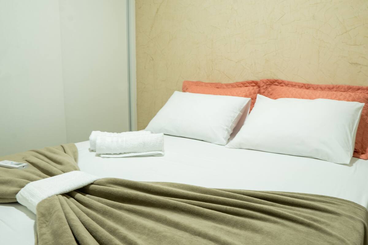 Un lit bien fait, avec des oreillers blancs et orange, une couverture beige et une télécommande posée sur le côté.