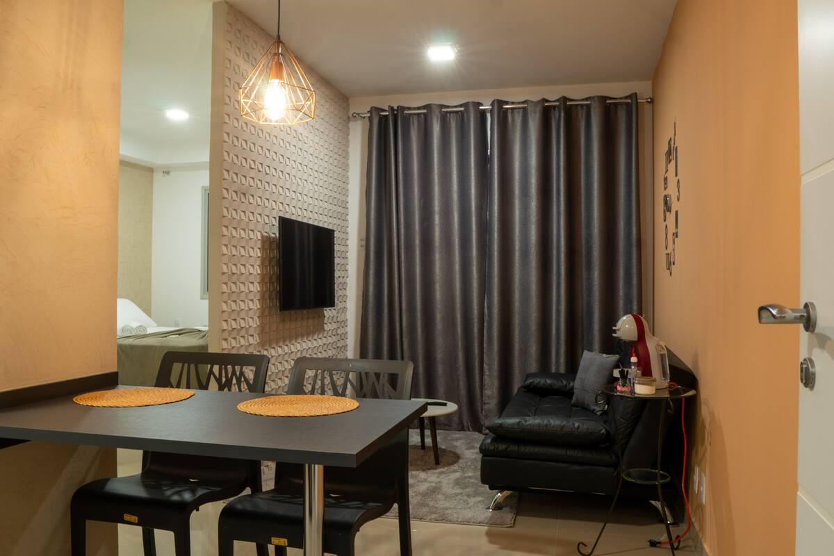 Un espace de vie compact comprenant un coin repas, un canapé en cuir noir, un téléviseur fixé à un mur texturé et une fenêtre recouverte d’un rideau.