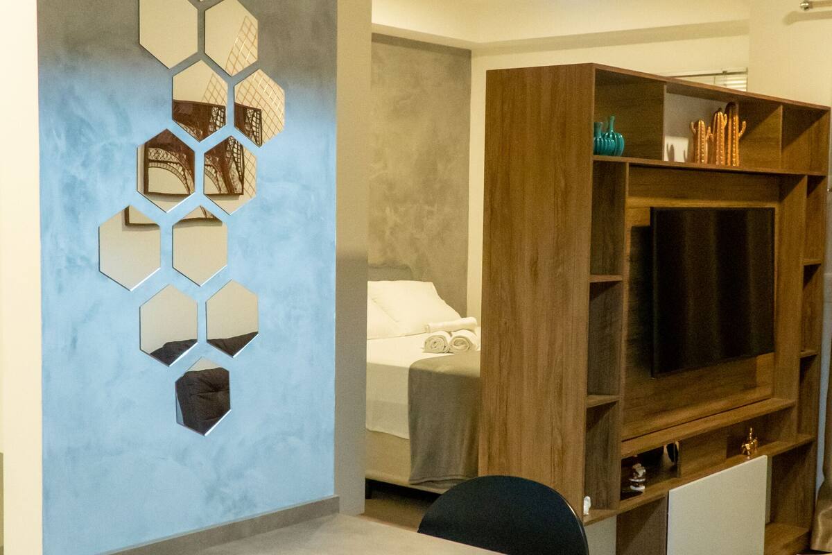 Un espace repas moderne avec une longue table, des chaises, un mur décoré d’un grand miroir hexagonal et un meuble de rangement en bois intégrant un téléviseur à écran plat.