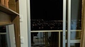 Quarto com porta de correr de vidro que dá para uma varanda com vista para a cidade à noite.