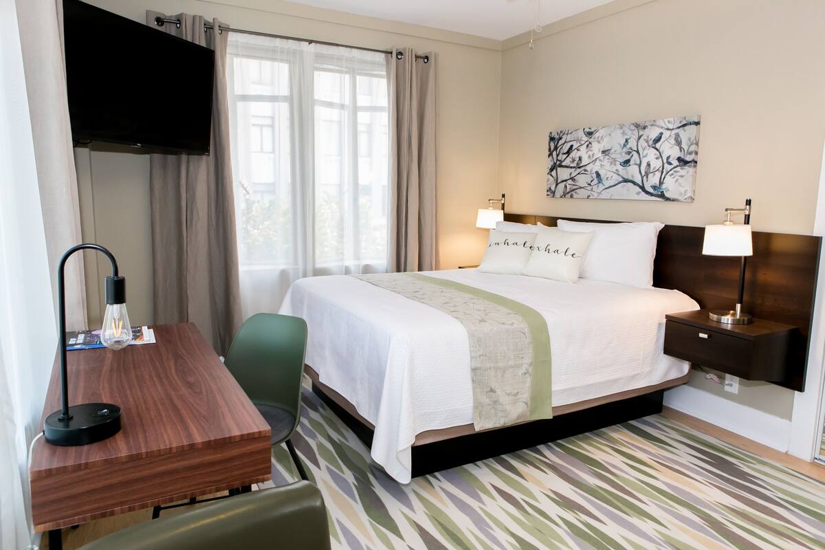 A hotel room with a bed, bedside lamps, a desk with a lamp, a chair, and a painting on the wall.