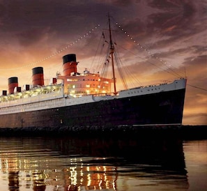 A large, historic ship with multiple decks and smokestacks, moored at a dock with a sunset in the background. - Hotel Metropolitan (Long Beach)