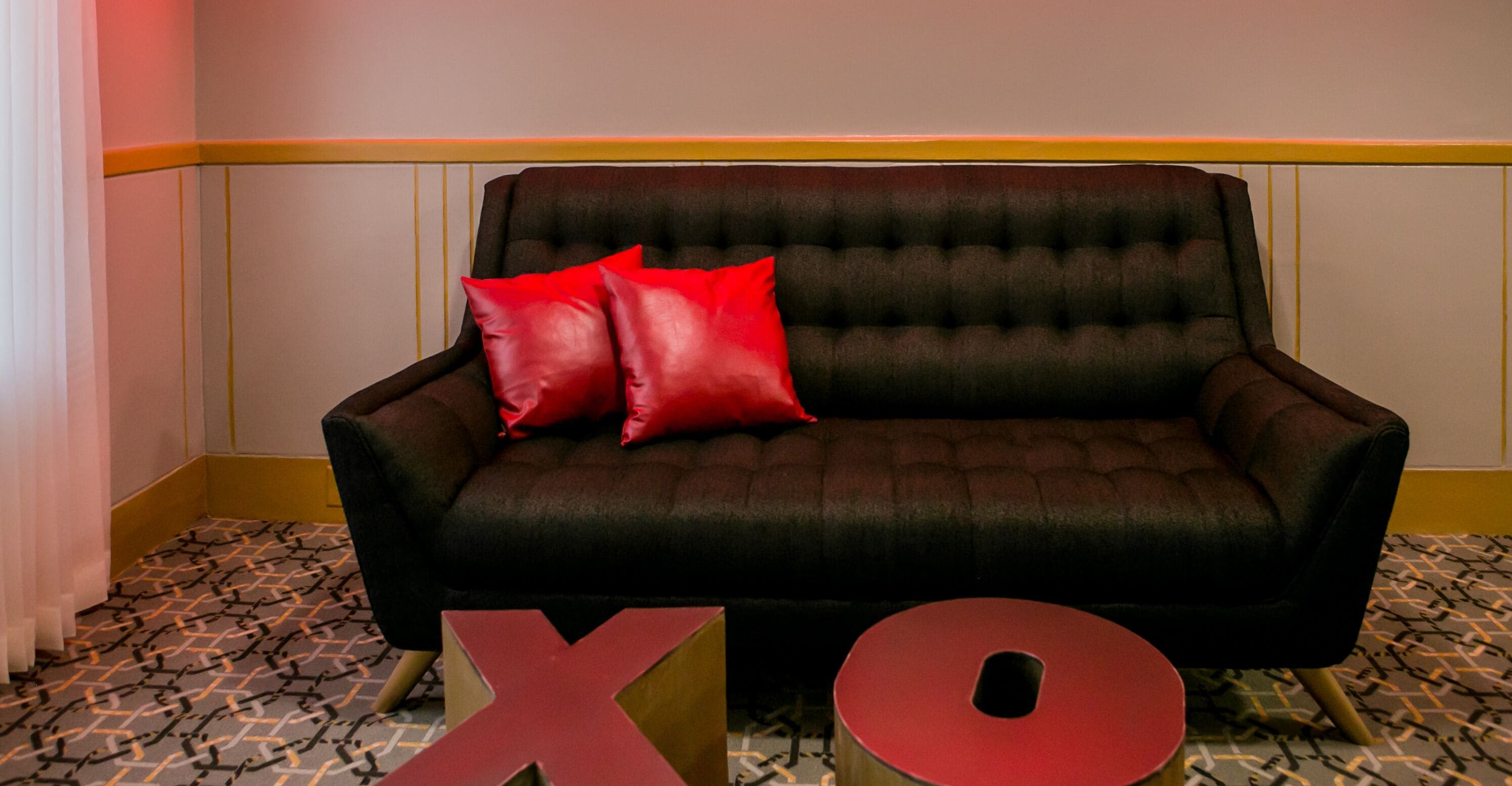 Un sofá negro con cojines rojos en una habitación con suelo estampado y una franja amarilla en la pared.