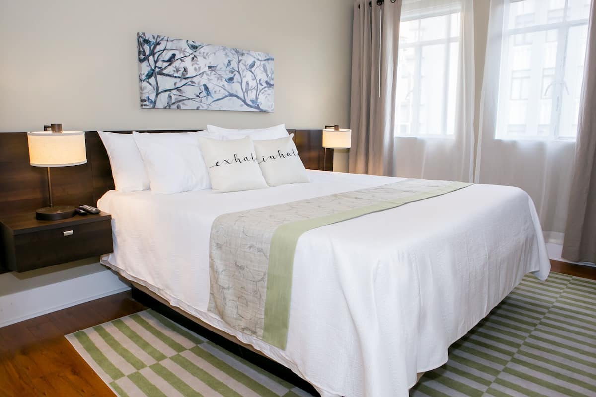 A neatly made bed with white bedding and a green accent, a nightstand with a lamp, and a wall-mounted artwork above the bed.