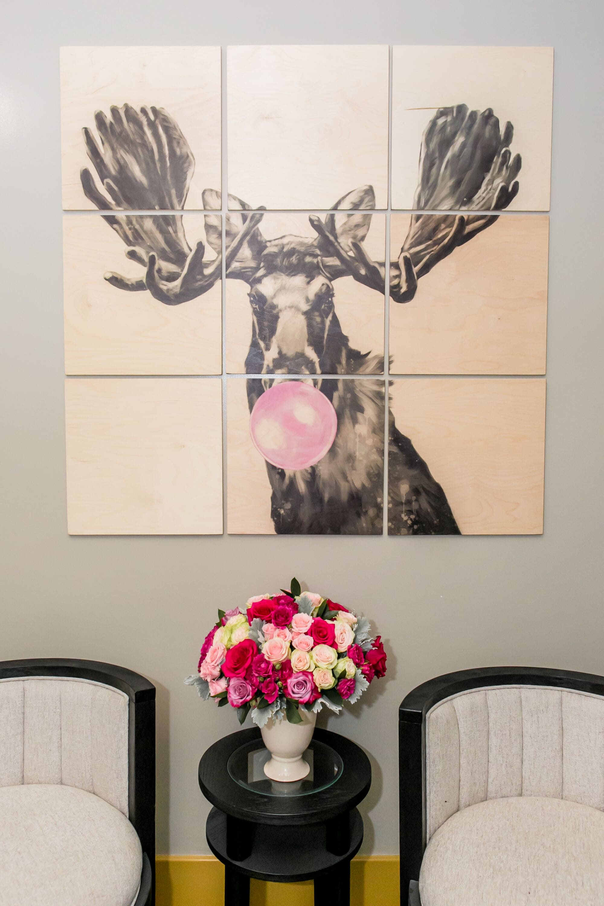Une œuvre d’art murale représentant un élan avec ses bois et un ballon rose, encadrée de deux chaises et d’une petite table sur laquelle est posé un bouquet de fleurs.