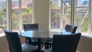 Una habitación con una mesa de vidrio y cuatro sillas, ventanas amplias con vista a la ciudad, y piso de madera.