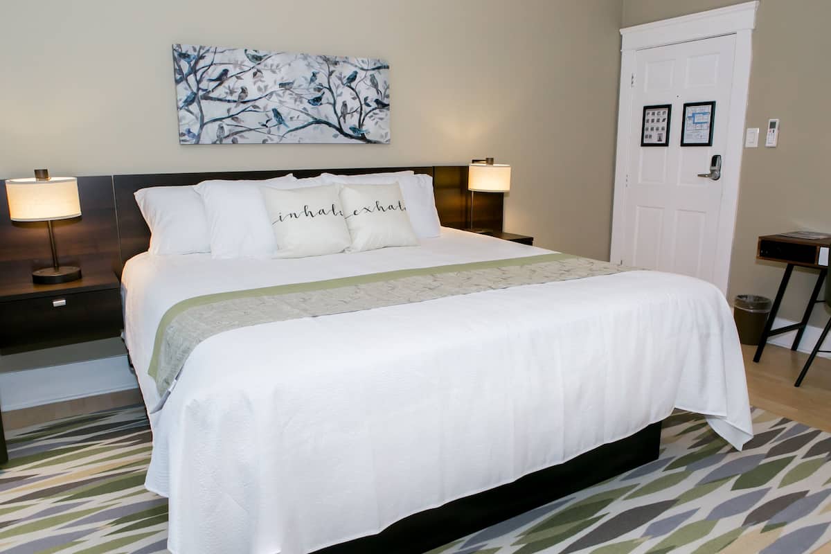 A neatly made bed with white linens and a green accent, flanked by two bedside lamps, in a room with a patterned rug and a wall-mounted abstract art piece.