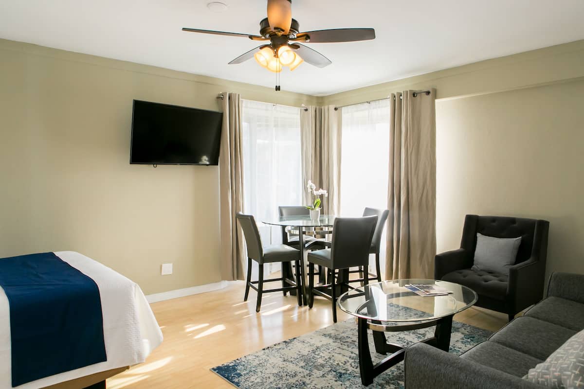 A hotel room with a bed, a sofa, a coffee table, a dining table with chairs, a ceiling fan, a flat-screen TV, and large windows with curtains.