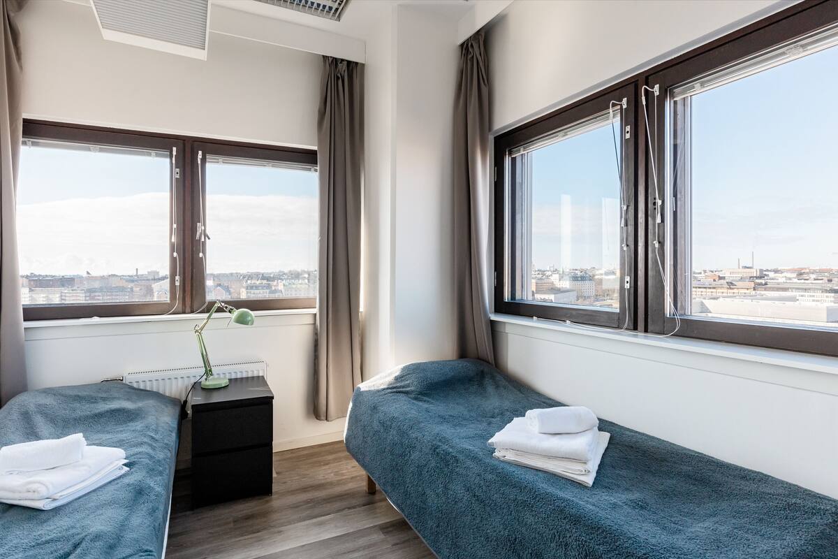 A hotel room with two single beds, a desk with a lamp, and large windows offering a city view.