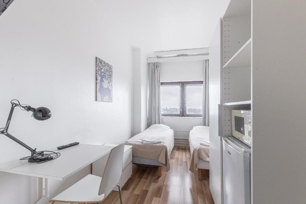 A small, modern room with a desk, two beds, a chair, a desk lamp, a window with curtains, and a microwave.