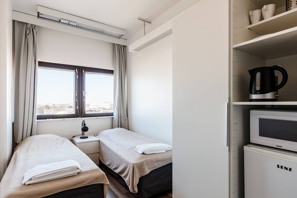 A hotel room with two beds, a window with curtains, a desk with a lamp, a kettle, and a microwave.