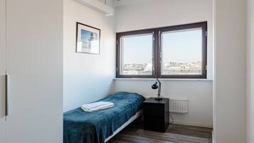 A small, tidy room with a single bed, a radiator, a window with a view of buildings, and a framed picture on the wall.