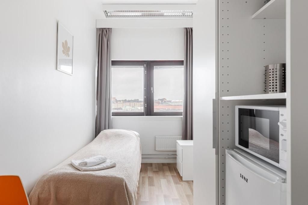 A small, modern room with a bed, a window with curtains, a radiator, and a kitchenette with a microwave.