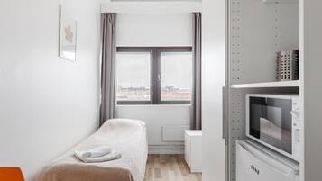 A small, modern room with a bed, a window with curtains, a radiator, and a kitchenette with a microwave.