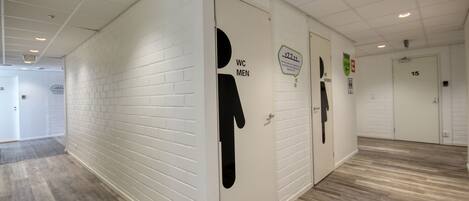 A hallway with white brick walls and wooden flooring, featuring doors labeled "WC MEN" and "WC WOMEN".
