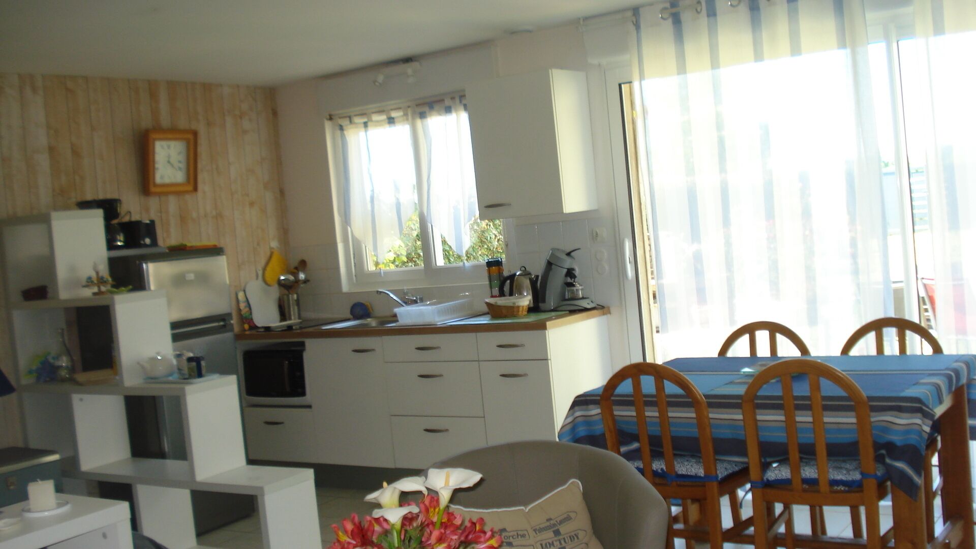 A compact kitchen with white cabinets, a sink, and a window with curtains. There is a dining area with a table and chairs, and a flower arrangement on the table.