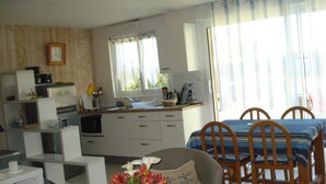 Una cocina compacta con armarios blancos, un lavamanos y una ventana con cortinas. Hay una zona de comedor con una mesa y sillas, y un arreglo floral sobre la mesa.