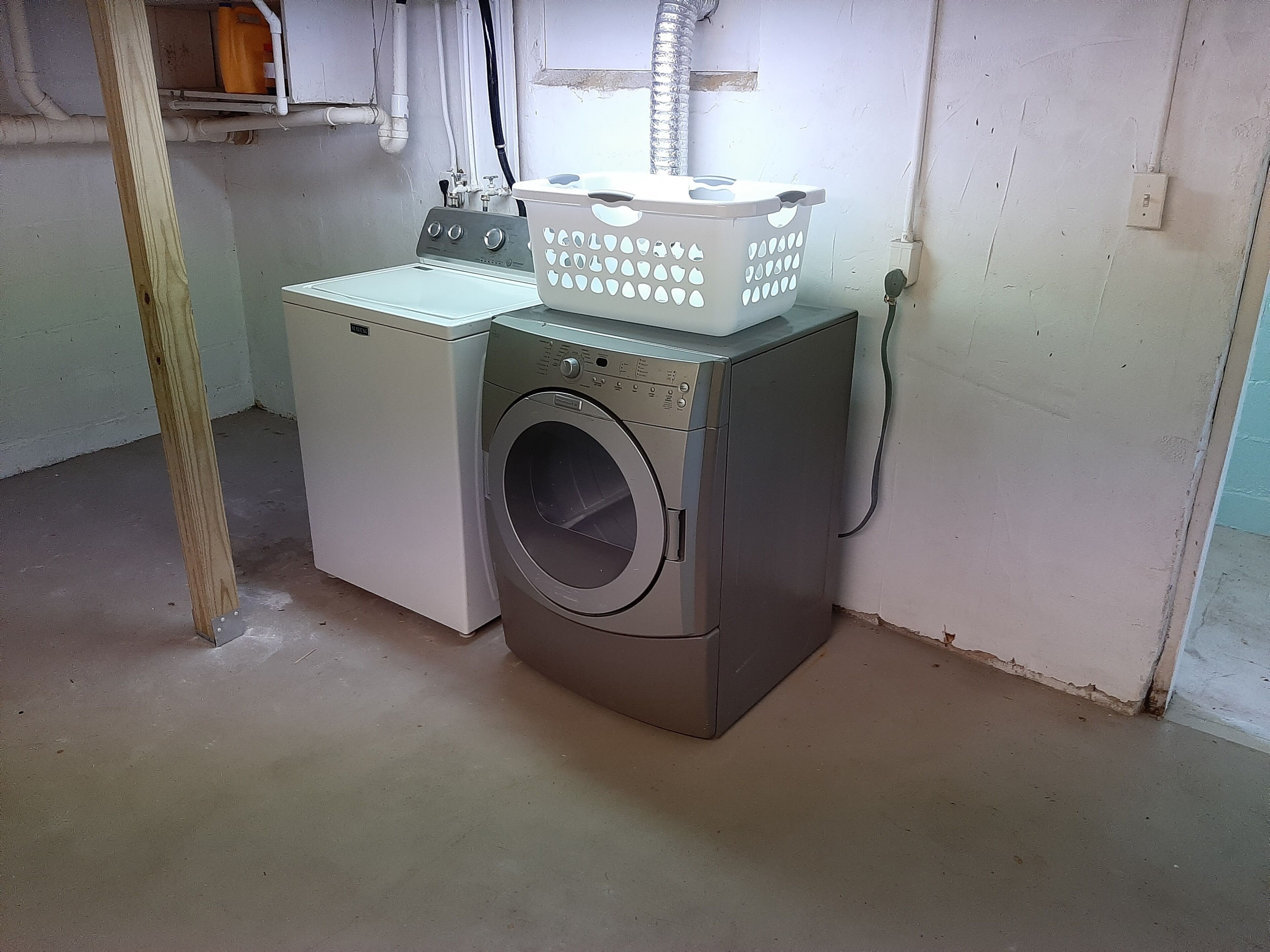 A laundry area with a washing machine and dryer, a plastic basket, and a power outlet on the wall.