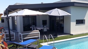 Un espace piscine extérieur moderne, doté de chaises longues et de parasols, un coin repas avec tables et chaises, et un bâtiment avec de grandes fenêtres et un toit plat.