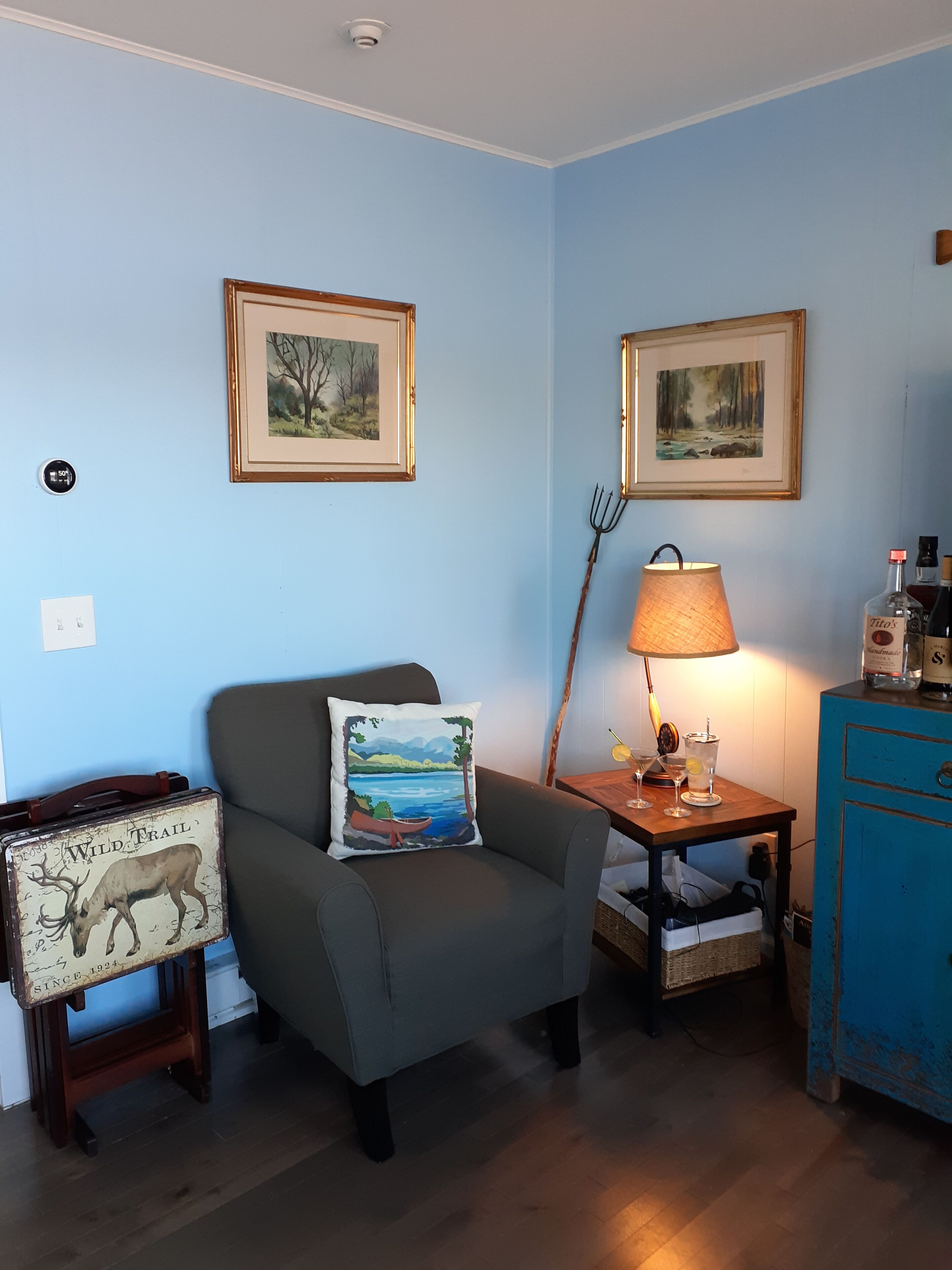 A living room with a grey armchair, a small table with a lamp, two framed paintings, and a blue cabinet.
