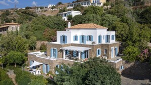 Una casa de varios pisos con terraza, rodeada de vegetación y otros edificios.
