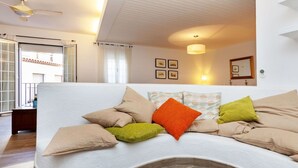 A modern living room with a curved white sofa, multiple cushions, a ceiling fan, and a large window with curtains. - Beautiful  villa for 5 guests with WIFI, TV and balcony (Tossa De Mar)