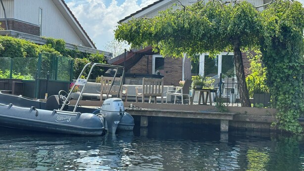 Ein Boot liegt an einem Haus mit Garten und überdachter Terrasse festgemacht.