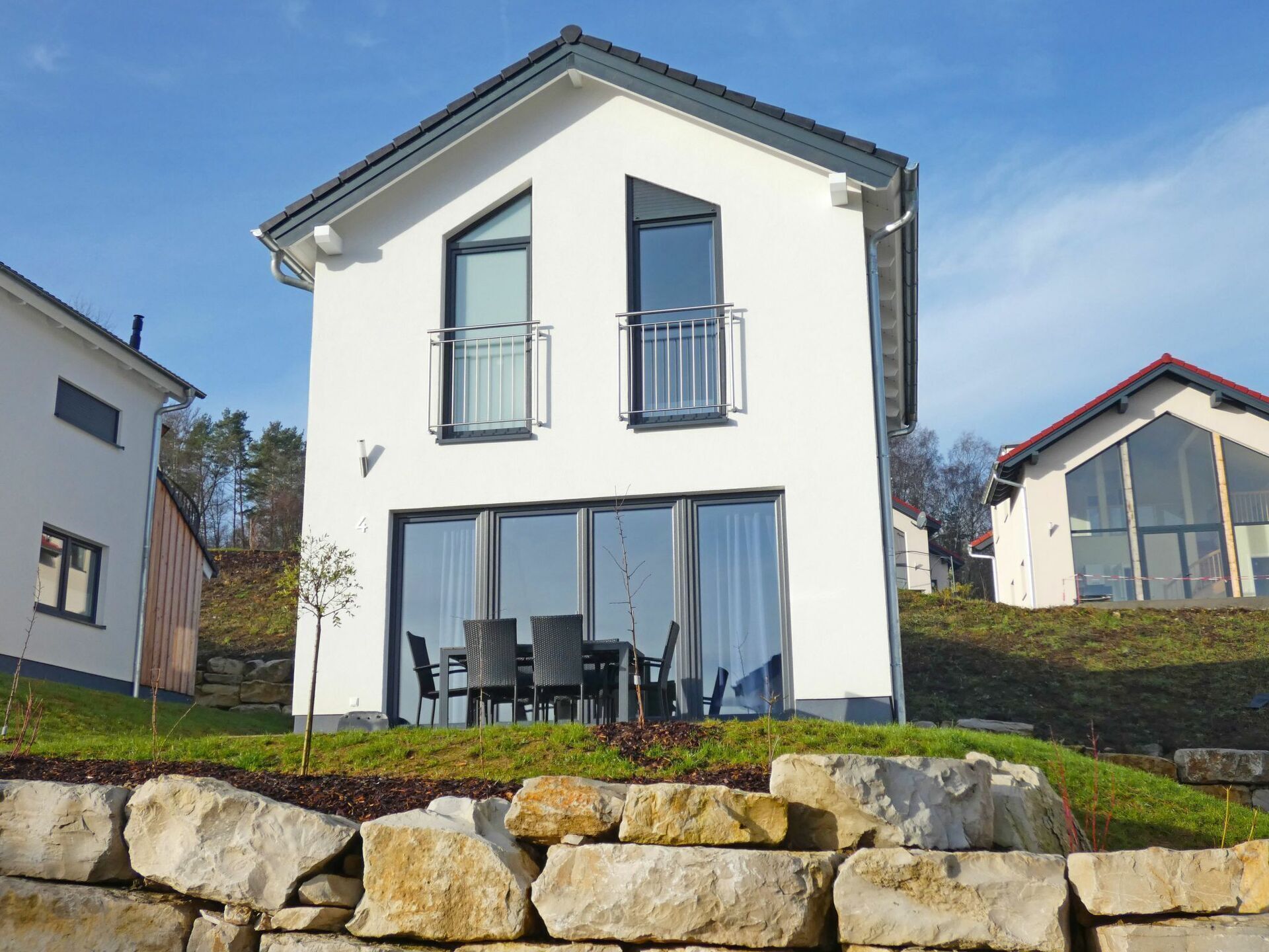 Ein modernes, zweistöckiges Haus mit Balkon, großen Fenstern und einer Steinmauer im Vordergrund.